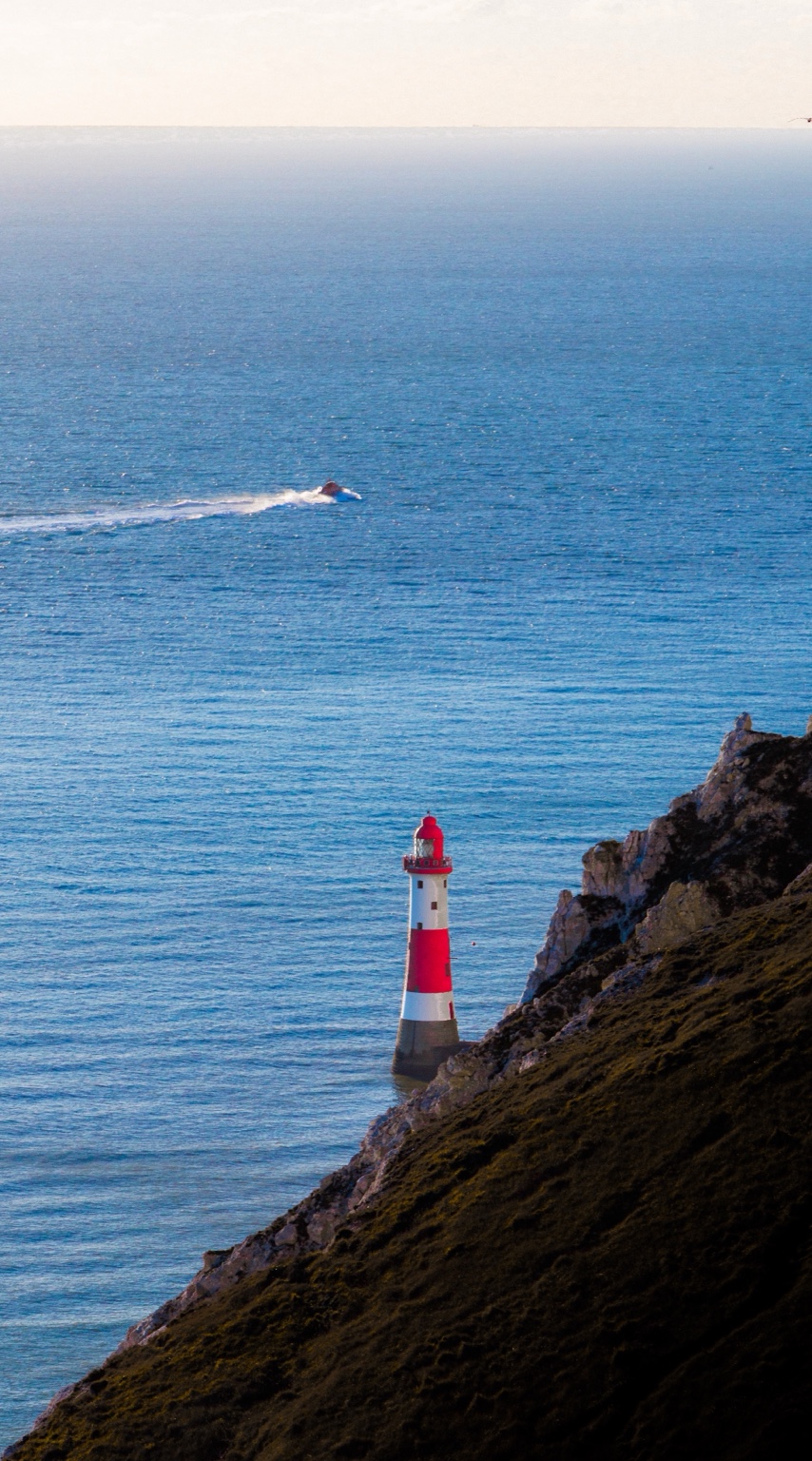 A red and white lighthouse beneath a cliff overlooking blue sea.